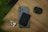 A smartphone with a black wallet attached to its back is placed on a wooden surface. Next to the smartphone are a pair of white wireless earbuds and a black carrying case. There is a hint of green foliage in the top left corner.