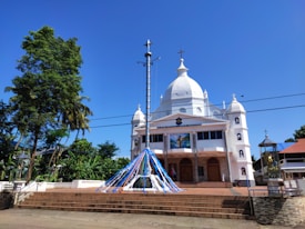 A large white church with multiple domes and crosses is set against a clear blue sky. It is surrounded by lush green trees and plants. In front of the church, an open area with steps leads up to the entrance, which is adorned with colorful ribbons. Statues and religious icons are visible on the facade.