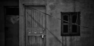 A locked wooden door with ornate circular designs on it is situated next to a small window with double panes. The structure appears weathered, with visible cracks and stains on the wall. A taut wire is stretched diagonally across the facade.