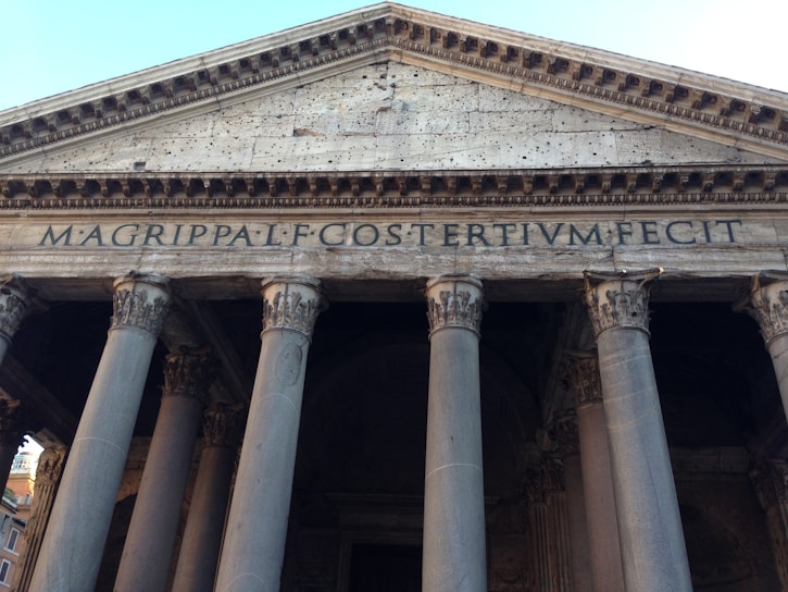 The image captures the front facade of an ancient building with a prominent triangular pediment supported by large Corinthian columns. The stone is weathered and inscribed with Latin text that reads 'MAGRIPPA L F COSTERTIVM FECIT.'