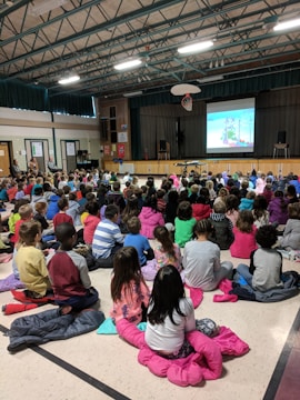 A large group of children sitting on the floor in a school gymnasium, watching a projected video on a screen at the front. The ceiling features exposed beams and fluorescent lights. A basketball hoop is visible above the stage. The children are wearing colorful clothing, and some sit on jackets or blankets.