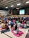 A large group of children sitting on the floor in a school gymnasium, watching a projected video on a screen at the front. The ceiling features exposed beams and fluorescent lights. A basketball hoop is visible above the stage. The children are wearing colorful clothing, and some sit on jackets or blankets.