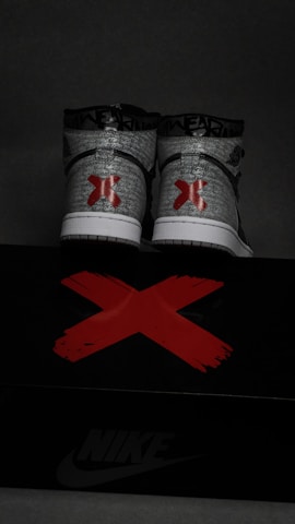 A pair of high-top sneakers with a graffiti-like gray and white pattern, featuring bold red 'X' designs on the back. The sneakers are placed on a black shoe box marked with a large red 'X' and the Nike logo in white.