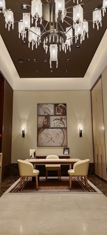 A well-furnished and elegant office space featuring a modern chandelier with crystal elements hanging from a ceiling, illuminating a cozy meeting area with a wooden table surrounded by three cream-colored chairs. The back wall displays an art piece with vintage clock motifs, flanked by two wall sconces.