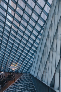 A modern architectural interior featuring a glass and metal framework ceiling. The geometric pattern is formed by intersecting lines creating a grid-like structure. Below, there are rows of tables and chairs, possibly indicating a study area or library. The lighting is subtle, with some lamps providing illumination, adding a cozy contrast to the industrial design.