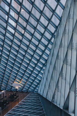 A modern architectural interior featuring a glass and metal framework ceiling. The geometric pattern is formed by intersecting lines creating a grid-like structure. Below, there are rows of tables and chairs, possibly indicating a study area or library. The lighting is subtle, with some lamps providing illumination, adding a cozy contrast to the industrial design.