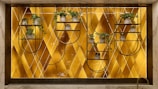 A decorative wall features a geometric patterned background with a warm golden hue. Mounted metal frames hold several small potted plants with lush green foliage, arranged symmetrically. The design blends natural elements with modern art deco style.