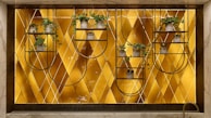 A decorative wall features a geometric patterned background with a warm golden hue. Mounted metal frames hold several small potted plants with lush green foliage, arranged symmetrically. The design blends natural elements with modern art deco style.