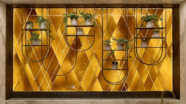 A decorative wall features a geometric patterned background with a warm golden hue. Mounted metal frames hold several small potted plants with lush green foliage, arranged symmetrically. The design blends natural elements with modern art deco style.