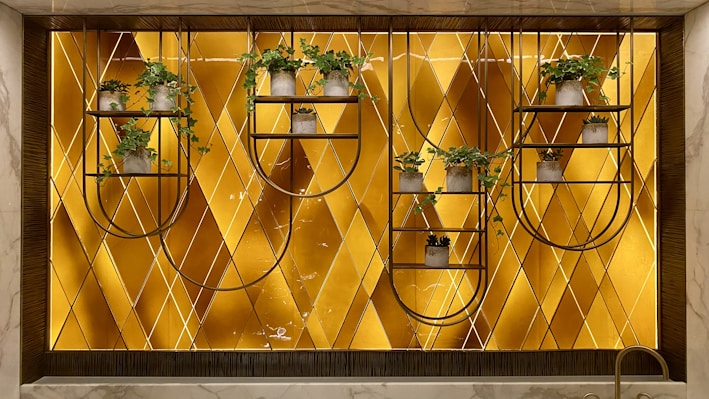 A decorative wall features a geometric patterned background with a warm golden hue. Mounted metal frames hold several small potted plants with lush green foliage, arranged symmetrically. The design blends natural elements with modern art deco style.