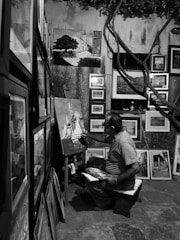 A person is seated in a small, dimly lit room surrounded by numerous framed paintings. They are actively painting on a canvas with a variety of art supplies around them. The setting suggests a cozy, creative space with artworks of different sizes displayed on the walls and leaning against surfaces.