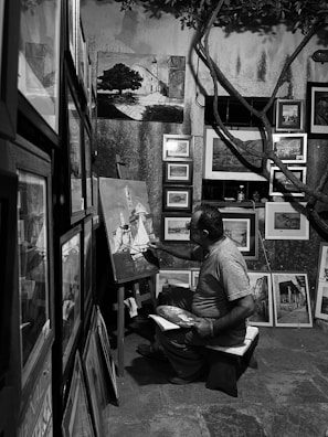 A person is seated in a small, dimly lit room surrounded by numerous framed paintings. They are actively painting on a canvas with a variety of art supplies around them. The setting suggests a cozy, creative space with artworks of different sizes displayed on the walls and leaning against surfaces.