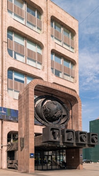 A tall building with a unique architectural design featuring curved windows and a stone facade. Prominently displayed is a large emblem with a globe and Cyrillic text, situated above a modern entrance. The structure is bathed in natural light against a backdrop of a clear blue sky.