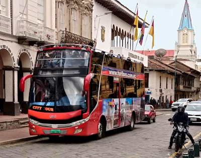 A double-decker tour bus with an open upper deck is traveling down a street lined with historic buildings. There are several flags waving atop one of the buildings, and a church with a blue and white steeple is visible in the background. People are seen both on the bus and walking on the sidewalk. A motorcyclist is positioned in the foreground, and other vehicles are present on the street.