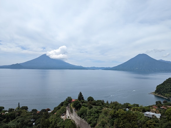 A serene lake is bordered by lush green vegetation and framed by two prominent volcanoes under a partly cloudy sky. There are some buildings and roads nestled among the dense greenery along the shoreline, with gentle waves visible on the lake's surface.