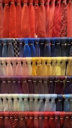 A collection of neatly arranged neckties organized by color, featuring various patterns such as stripes, polka dots, and checks. The display includes a diverse range of colors with each row showcasing a different color palette. The ties hang in orderly rows, creating a visually appealing and colorful composition.