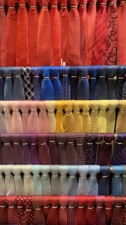 A collection of neatly arranged neckties organized by color, featuring various patterns such as stripes, polka dots, and checks. The display includes a diverse range of colors with each row showcasing a different color palette. The ties hang in orderly rows, creating a visually appealing and colorful composition.