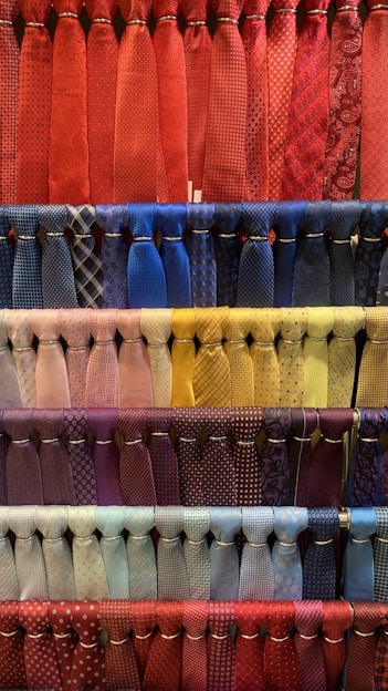 A collection of neatly arranged neckties organized by color, featuring various patterns such as stripes, polka dots, and checks. The display includes a diverse range of colors with each row showcasing a different color palette. The ties hang in orderly rows, creating a visually appealing and colorful composition.