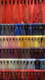 A collection of neatly arranged neckties organized by color, featuring various patterns such as stripes, polka dots, and checks. The display includes a diverse range of colors with each row showcasing a different color palette. The ties hang in orderly rows, creating a visually appealing and colorful composition.