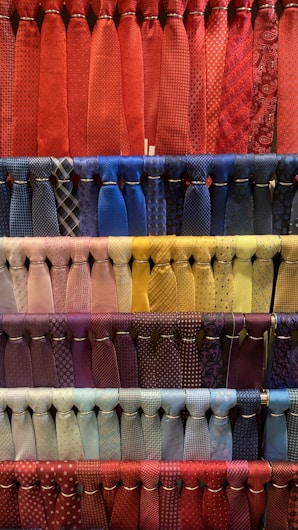 A collection of neatly arranged neckties organized by color, featuring various patterns such as stripes, polka dots, and checks. The display includes a diverse range of colors with each row showcasing a different color palette. The ties hang in orderly rows, creating a visually appealing and colorful composition.