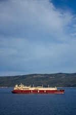 A large LNG (liquefied natural gas) ship is sailing on a calm blue sea with a backdrop of lush, green hills under a partly cloudy sky.