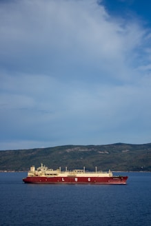 A large LNG (liquefied natural gas) ship is sailing on a calm blue sea with a backdrop of lush, green hills under a partly cloudy sky.