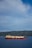 A large LNG (liquefied natural gas) ship is sailing on a calm blue sea with a backdrop of lush, green hills under a partly cloudy sky.