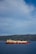 A large LNG (liquefied natural gas) ship is sailing on a calm blue sea with a backdrop of lush, green hills under a partly cloudy sky.