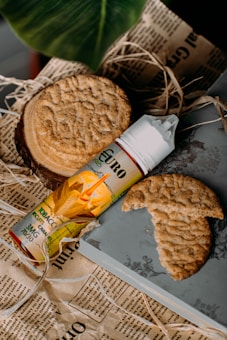 An arrangement featuring a round, textured cookie resting on a slice of wood, next to a cylindrical bottle labeled 'Euro' and adorned with an image of corn and the text 'Tobacco'. Another cookie is partially broken on a gray surface. The items are placed on printed newspaper-like paper and surrounded by a few straw-like elements, with a large, green leaf partially visible in the background.