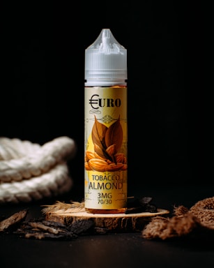 A bottle labeled 'Euro Tobacco Almond 3MG 70/30' with an illustration of almonds and tobacco leaves. It stands on a textured wooden surface surrounded by natural materials like rope, wood chips, and fibers against a dark background.
