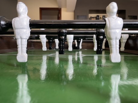Several foosball players are attached to rods on a foosball table, arranged in an alternating pattern of white and black pieces. The field below has a glossy green surface, indicating a soccer pitch layout. The background is slightly out of focus, featuring some furniture and a doorway.