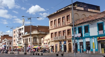 A vibrant street scene featuring several historic buildings with ornate architectural details, colorful facades, and tiled roofs. A busy pedestrian area with numerous people walking and small market stalls under multi-colored umbrellas. The sky is clear with a few fluffy clouds, and street lamps are visible along the pavement.