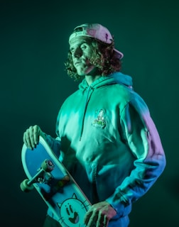 A person wearing a hoodie and a backwards cap holds a skateboard with a cartoon graphic. The lighting is a mix of green and teal, creating a vibrant and edgy vibe.