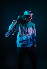 A person is wearing a hoodie and a baseball cap, holding a skateboard over their shoulder. The lighting creates a moody ambiance with teal and dark tones. The person looks relaxed and casual.