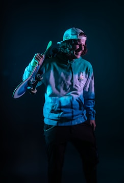 A person is wearing a hoodie and a baseball cap, holding a skateboard over their shoulder. The lighting creates a moody ambiance with teal and dark tones. The person looks relaxed and casual.