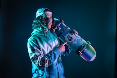 A person wearing a cap and a tie-dye hoodie is holding a colorful skateboard decorated with a cartoon ghost illustration. The lighting casts a blue tint across the scene, creating a vibrant and playful atmosphere.