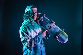 A person wearing a cap and a tie-dye hoodie is holding a colorful skateboard decorated with a cartoon ghost illustration. The lighting casts a blue tint across the scene, creating a vibrant and playful atmosphere.