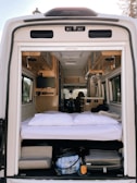 The interior of a camper van is shown from the open back doors, featuring a cozy sleeping area with white bedding and pillows. Above the bed, the van interior includes wooden cabinets and a small television mounted on the side. The space under the bed is used for storage, containing various items such as a suitcase and other travel gear. The materials and finishes suggest a well-organized and functional living area suitable for travel.