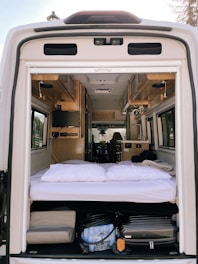The interior of a camper van is shown from the open back doors, featuring a cozy sleeping area with white bedding and pillows. Above the bed, the van interior includes wooden cabinets and a small television mounted on the side. The space under the bed is used for storage, containing various items such as a suitcase and other travel gear. The materials and finishes suggest a well-organized and functional living area suitable for travel.
