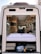 The interior of a camper van is shown from the open back doors, featuring a cozy sleeping area with white bedding and pillows. Above the bed, the van interior includes wooden cabinets and a small television mounted on the side. The space under the bed is used for storage, containing various items such as a suitcase and other travel gear. The materials and finishes suggest a well-organized and functional living area suitable for travel.