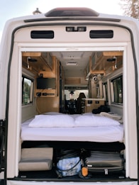The interior of a camper van is shown from the open back doors, featuring a cozy sleeping area with white bedding and pillows. Above the bed, the van interior includes wooden cabinets and a small television mounted on the side. The space under the bed is used for storage, containing various items such as a suitcase and other travel gear. The materials and finishes suggest a well-organized and functional living area suitable for travel.