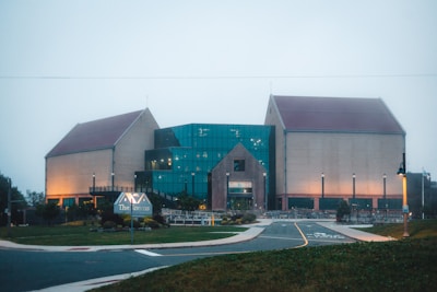 A large building with distinct architecture features a central section made of glass and flanked by two brick structures with sloped roofs. The area is well-lit with street lamps, and there is a sign in the foreground. The surrounding environment includes a paved road, grass, and neatly arranged plants.