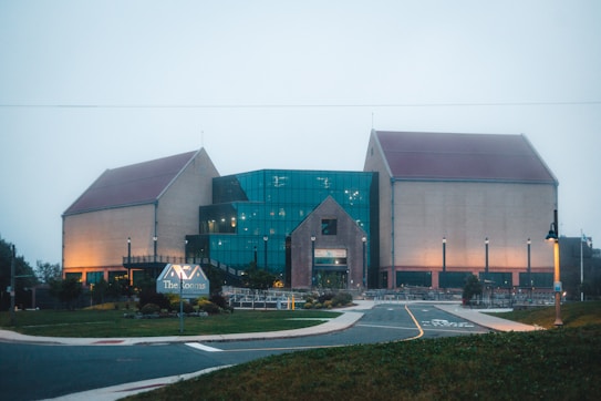 A large building with distinct architecture features a central section made of glass and flanked by two brick structures with sloped roofs. The area is well-lit with street lamps, and there is a sign in the foreground. The surrounding environment includes a paved road, grass, and neatly arranged plants.