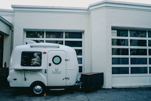 A small, white camper is parked in front of a building with large, garage-style windows. The camper has the words 'Bearcat Lounge' written on the side and features a small circular window on the door.