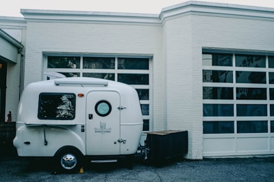 A small, white camper is parked in front of a building with large, garage-style windows. The camper has the words 'Bearcat Lounge' written on the side and features a small circular window on the door.