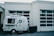 A small, white camper is parked in front of a building with large, garage-style windows. The camper has the words 'Bearcat Lounge' written on the side and features a small circular window on the door.