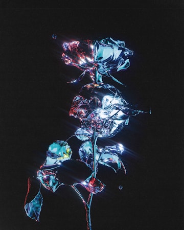 A digitally manipulated image of a rose and leaves appearing to be made of reflective, metallic material. The surface glistens with a mixture of blue, red, and silver hues, set against a dark background. Light reflections and lens flares add a futuristic, ethereal quality to the scene.