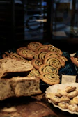 A variety of baked goods are displayed, including a pile of spiraled pastries with green filling, identified by a sign as 'Escargot Chocolat Pistache.' Additionally, there is sliced, rustic-looking bread and a small dish containing pastries.