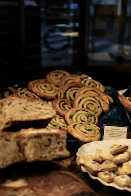 A variety of baked goods are displayed, including a pile of spiraled pastries with green filling, identified by a sign as 'Escargot Chocolat Pistache.' Additionally, there is sliced, rustic-looking bread and a small dish containing pastries.