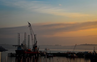 A peaceful harbor scene at sunset with an offshore rig silhouetted against a colorful sky. Cranes and the rig’s tall structures are prominent against the soft oranges and blues of the sunset. Calm water reflects the muted colors of the sky, and faint outlines of distant boats and land are visible.